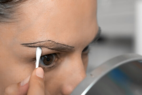 Young Woman Erases Contour Line Around Eyebrows With A Cotton Swab Before Painting Looking At Mirror, Closeup. Preparing To Eyebrow Paint Beauty Procedure For Herself At Home. Female Without Makeup.