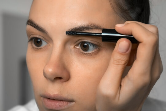 Woman is applying fixing gel on eyebrows using brush looking at mirror, closeup face. Beauty procedure at home. Modern trendy styling, correction, coloring of eyebrows.