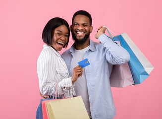 Cheerful black couple with credit card and shopping bags, buying gifts for Valentine's Day remotely on pink background