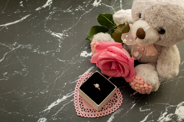 Cute teddy bear with pink rose and diamond ring on a black marble background.
