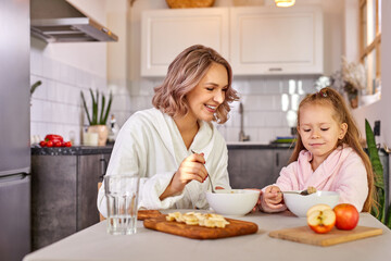 Obraz na płótnie Canvas girl enjoy meal for breakfast with mom, they sit together in modern kitchen at home, wearing domestic clothes, have meal