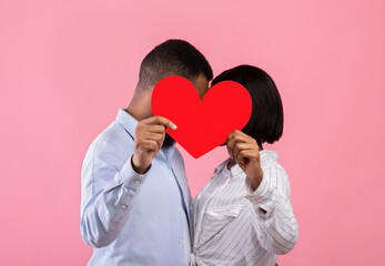 Romantic sweethearts hiding behind red paper heart and kissing on pink studio background