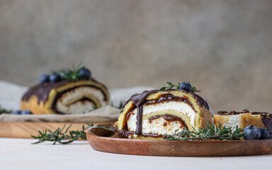Sponge cake roll with chocolate and cream cheese decorated with chocolate glaze, blueberry and rosemary on parchment, light concrete background. Biscuit swiss roll.