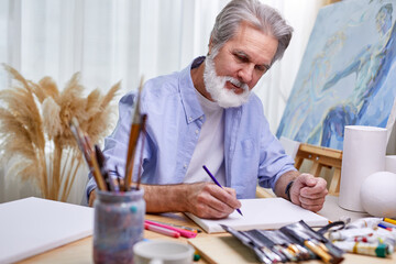 painter is drawing at home in light room, gray bearded man is creating a masterpiece using a pencil