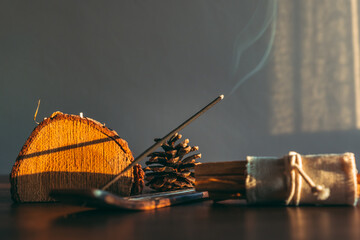 Burning incense stick in a scene natural and wooden decoration objects during sunset. Selective focus on stick tip ash. Cozy home environment. - obrazy, fototapety, plakaty