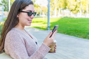 Side view of a smiling woman using a smart phone with coffee to go sitting down in a park.