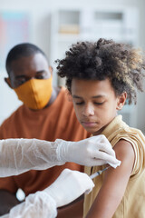 Vertical portrait of unrecognizable nurse vaccinating African-American boy against covid in clinic...