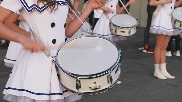 Young ladies in white marine short dresses play drums on a parade on summer day. Girls musicians drummers from municipal orchestra performing on festival. Close-up of hands with wooden sticks.