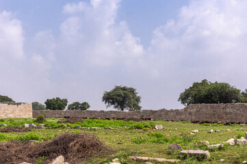 Hampi, Karnataka, India - November 5, 2013: Ancient Aqueduct. brown stone section of royal enclosure wall nearby under blue cloudscape. Green foliage and grass.