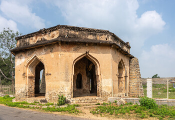 Fototapeta premium Hampi, Karnataka, India - November 5, 2013: Ancient Aqueduct. octagonal green brown stone ruinous water pavilion nearby under blue cloudscape. Green foliage and weeds around.