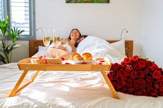 Romantic breakfast in bed on Valentine Day. Couple playing sexual game and having fun in bed celebrating holiday with champagne, gift, croissants and bouquet of red roses.