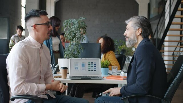 Businessmen mature and young are talking then doing high-five gand gesture and smiling looking at laptop screen with smartphone application design