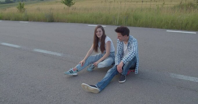 Couple of teenagers sitting in poses on a skateboard and having talk on road