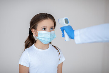 Little girl in face mask having her temperature checked by electronic thermometer while waiting for covid-19 vaccination