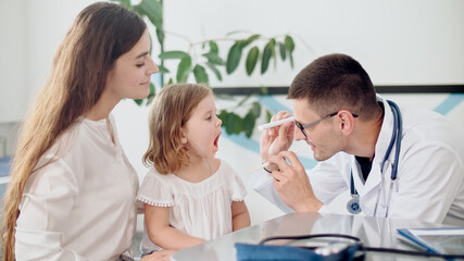 Fototapeta premium Male Doctor Pediatrician Examining an Ill Sad Kid Girl at Medical Visit With Mother in the Hospital. Male Family Doctor Examining and Consulting to Mother and Her Ill Child.