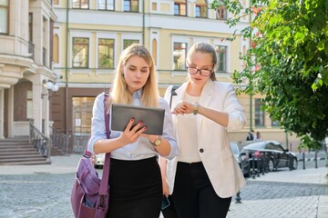 Two young businesswomen walking along city street, talking looking in digital tablet