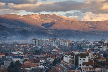 Misty, smoky cityscape of Pirot during sunset with impressive, rocky peak lighten by golden light in the background and a cloudy sky