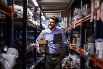 Fototapeta na wymiar Smiling bearded businessman walking trough storage of shipping firm, holding boxes under armpit and using tablet to check on goods.