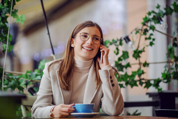 Young smiling brunette dressed smart casual sitting in cafe, holding cup of coffee and talking on the phone with a friend.