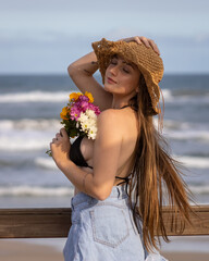 Balneário Gaivota, Santa Catarina, Brazil, November 2020. Model in front of the sea holding beautiful colorful flowers with a wavy brown hat illuminated by the sunlight.