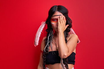 indian woman closing half of face isolated over red studio background, wearing shamanic clothes,...