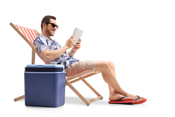Young man sitting in a deck chair next to a cooling box and holding a digital tablet