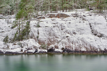 Fototapeta na wymiar A cliff with snow and trees with forest in the background and water in the foreground