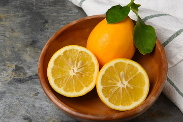 Closeup of a cut lemon and whole fruit with leaves in a wooden bowl on gray slate.