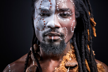 tribal guy with ethnic authentic make-up looks at camera, black african man with dreadlocks posing isolated over black background