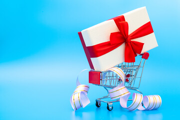Small shopping cart with gift box on blue background with festive ribbon. The concept of spending big money on the holidays. Copy space.