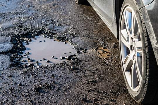 Car wheel close-up near a pothole on the road. Broken asphalt on the  roads. Spring and autumn problems for drivers. Potholes, water pits on a bad road.