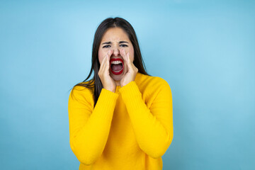 Young beautiful woman wearing yellow sweater over isolated blue background shouting and screaming loud to side with hands on mouth