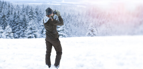A young hunter woman in the snowy forest. She watches wild animals in the forest using binoculars. Winter landscape. Banner photo.