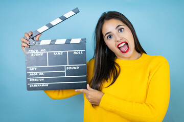 Young beautiful woman wearing yellow sweater over isolated blue background holding clapperboard...