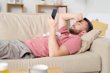 Young sick male patient using smartphone while lying on couch in living-room