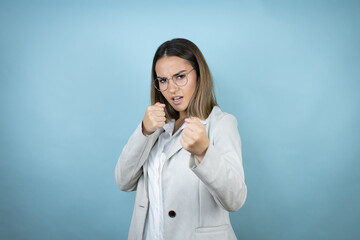 Young business woman over isolated blue background Punching fist to fight, aggressive and angry attack, threat and violence