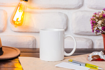 White mug cup is on the table with the background of a white brick wall and a lamp with yellow light.