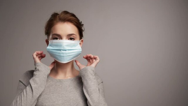 Woman putting on medical face protective mask, virus prevention..Portrait of young woman putting on protective medical mask, studio background. Real time 60fps. Social distancing and new normal.