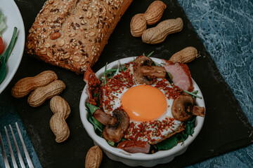 Perfectly cooked fried eggs with mushrooms, spices, pepper, meat and salt on arugula. Round breakfast plate on black plank with peanuts and baguette with seeds