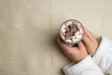 cappuccino in a transparent glass with marshmallows and cocoa powder in female hands on a textured beige fabric