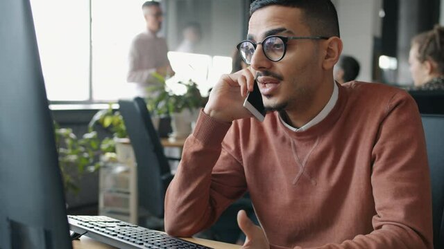 Serious Middle Eastern guy is calling on mobile phone and looking at computer screen working in office while colleagues are moving in background.