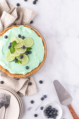 Top down view of a key lime pie garnished with lime, mint and blueberries against a light background.