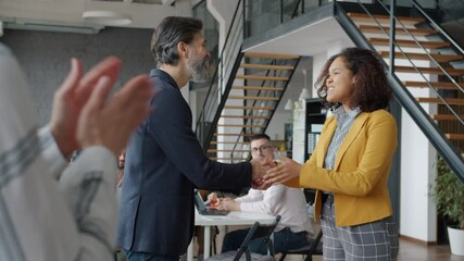 Boss mature man is shaking African American employee's hand giving good news while colleagues are clapping hands in office. Career and emotions concept.