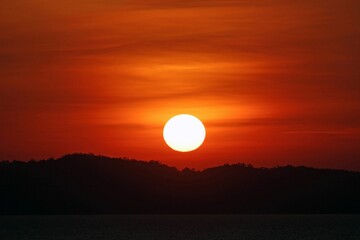 Full orb of the sun setting with the sky glowing orange over a silhouetted island and ocean, southeast Asia.
