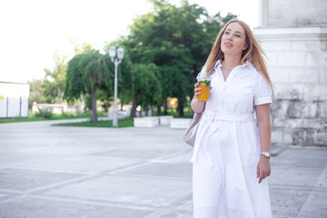 Beautiful overweight lady in white dress and holding the bagpack waking in the street of the city and smiling. Plus size  girl drink orange cocktail in the park
