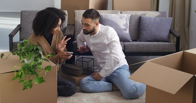Young family interracial couple surrounded by carton boxes looks at phone selects furniture online discusses solution looking for things in new apartment, concept of buying real estate and moving
