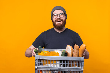 Cheerful young delivery man is holding a basket full of food and grocery over yellow background.