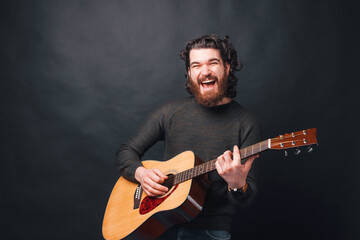 Joyful bearded man playing at acoustic guitar near dark background.