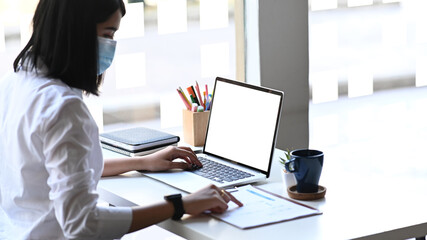 Fototapeta na wymiar Side view of businesswoman wearing protective mask working on laptop and analysing business report on white table.