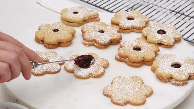 Flower shaped shortbread cookies filled with raspberry jam recipe. Woman hands using a kitchen knife spreading a layer of raspberry jam on flower shaped cookies